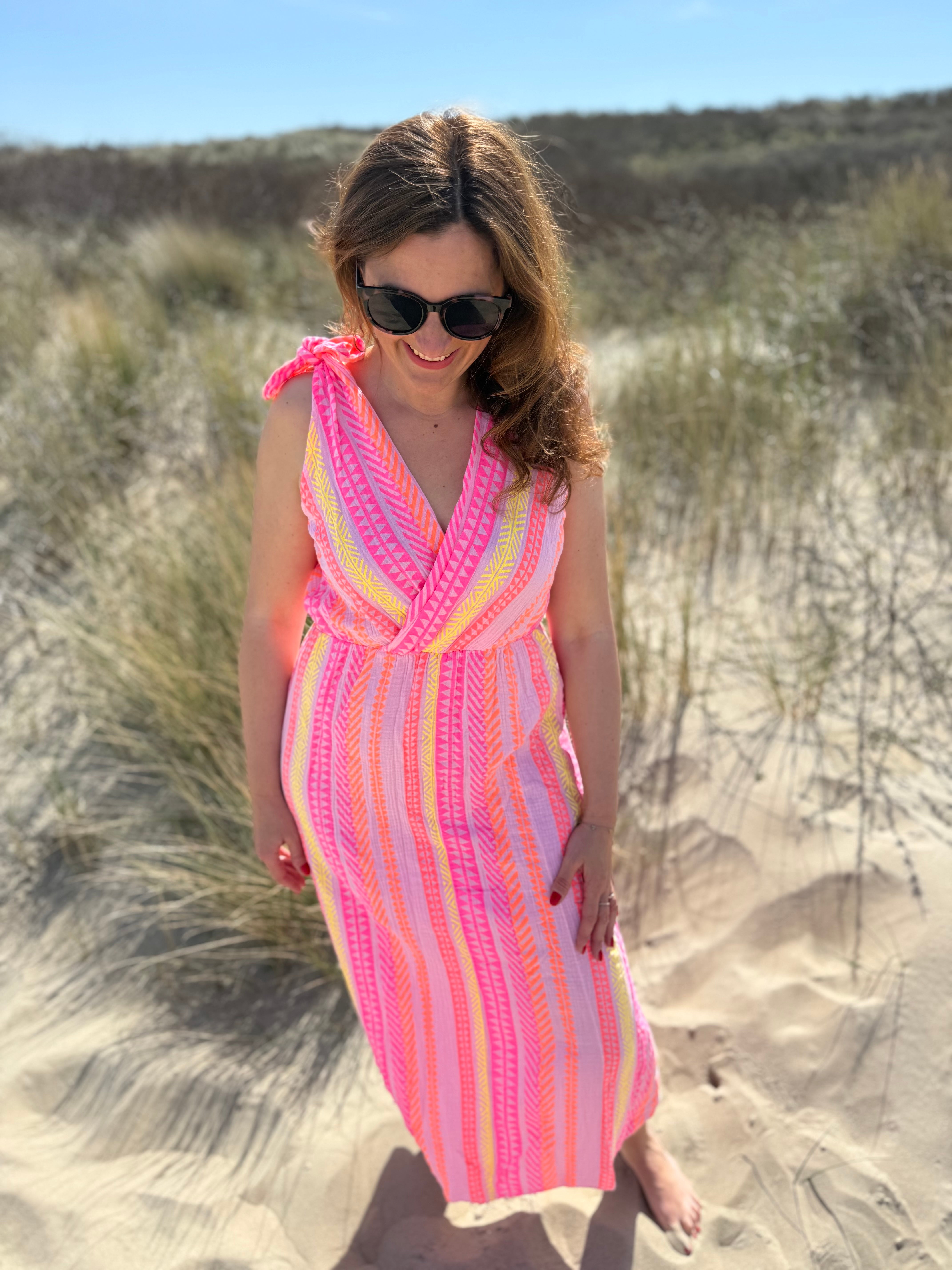 Musselinkleid in Midilänge mit V Ausschnitt. Es ist ärmellos und hat Schleifen an den Schultern zum binden. Das Kleid ist rosa mit pink, gelb und orangenen Ethno Muster.