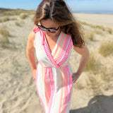 Musselinkleid in Midilänge mit V Ausschnitt. Es ist ärmellos und hat Schleifen an den Schultern zum binden. Das Kleid ist weiß mit pink und orangenen Ethno Muster.