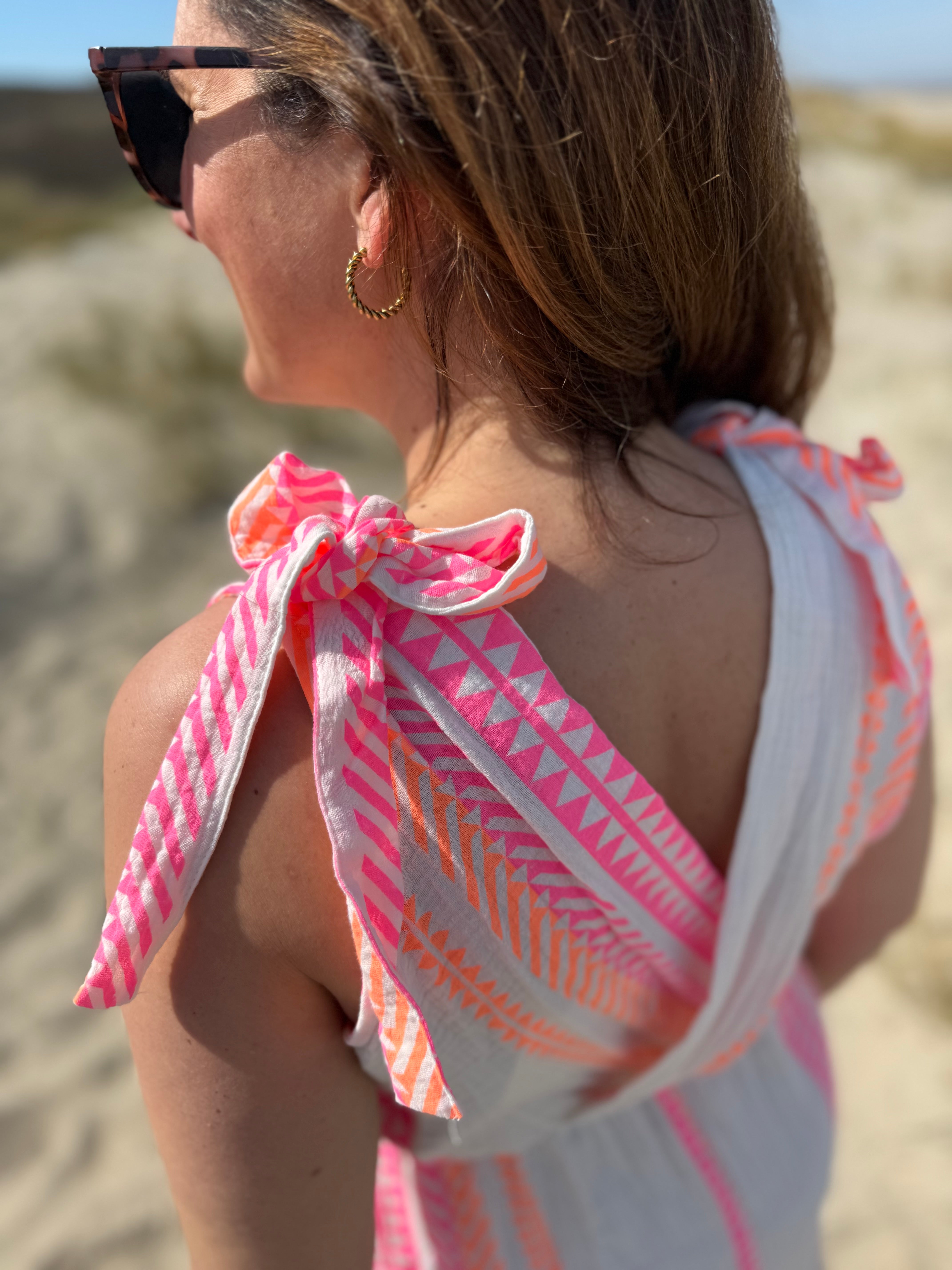 Musselinkleid in Midilänge mit V Ausschnitt. Es ist ärmellos und hat Schleifen an den Schultern zum binden. Das Kleid ist weiß mit pink und orangenen Ethno Muster.