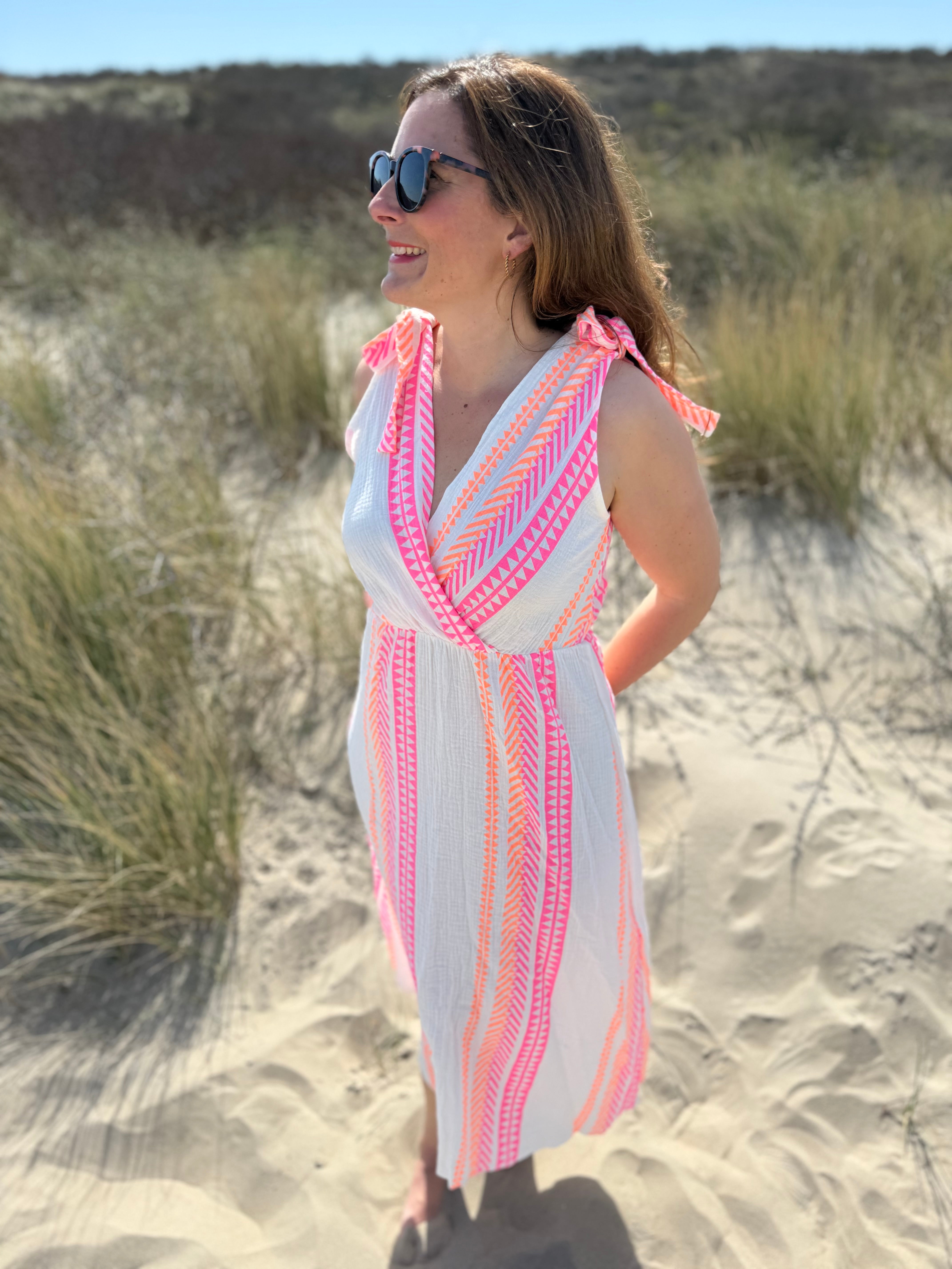 Musselinkleid in Midilänge mit V Ausschnitt. Es ist ärmellos und hat Schleifen an den Schultern zum binden. Das Kleid ist weiß mit pink und orangenen Ethno Muster.